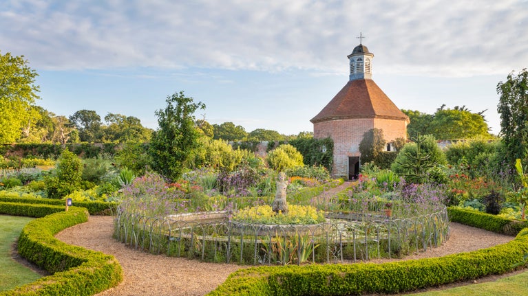 A circular pond, with a classical statue in the middle, in a formally designed garden with low green hedges lining the paths and borders filled with summer plants. At the back a brick-built hexagonal building with a cupola on top, and behind a line of trees